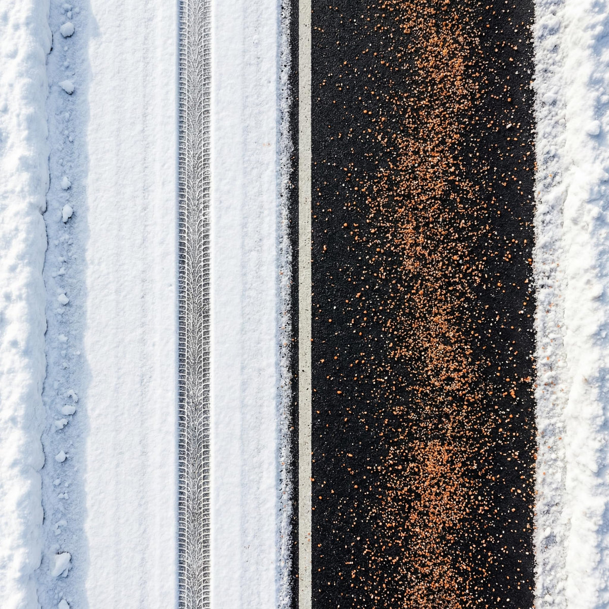 Overhead view of orange ice melt granules spread down a dark driveway between snow.