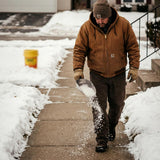 Man spreading salt from a scoop on a snowy walk with a yellow pail behind him.