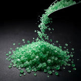 Green treated salt crystals pouring from a scoop onto a dark surface.