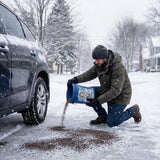 Man pouring Glacier Grip granules from a pail beside a car on a snowy street.
