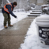 Worker in a safety vest scattering salt on a snowy sidewalk beside a pail.